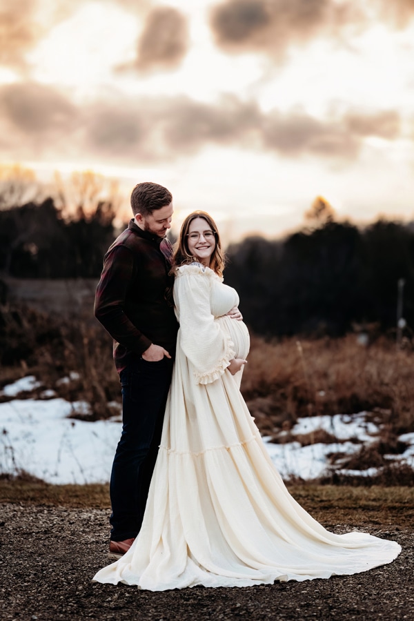 Family and newborn Photographer, a husband and expecting wife stand together outside, snow behind them in a meadow